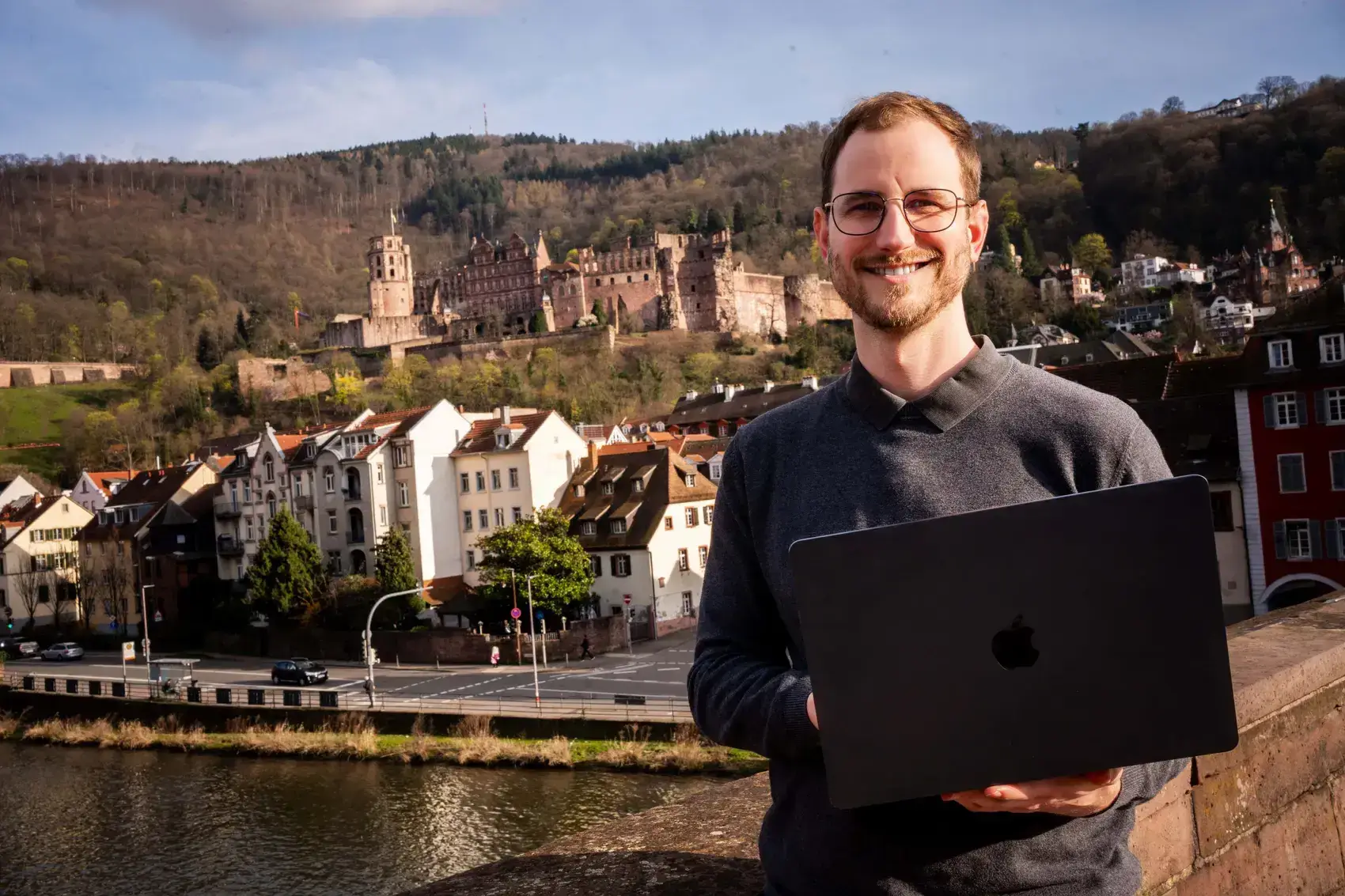 Joshua Stark mit Laptop an der Alten Brücke in Heidelberg, im Hintergrund das Heidelberger Schloss